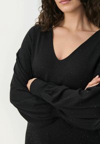 Fekete pulóver V-nyakkivágással és hosszú, bő ujjakkal. Apró, fényvisszaverő díszítésekkel ellátott textúrált minta jellemzi.