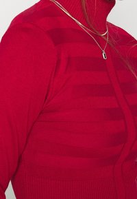 Pull à col roulé en maille rouge avec une texture rayée. Coupe ajustée avec des accents côtelés, mettant en valeur un tissu doux et chaud.