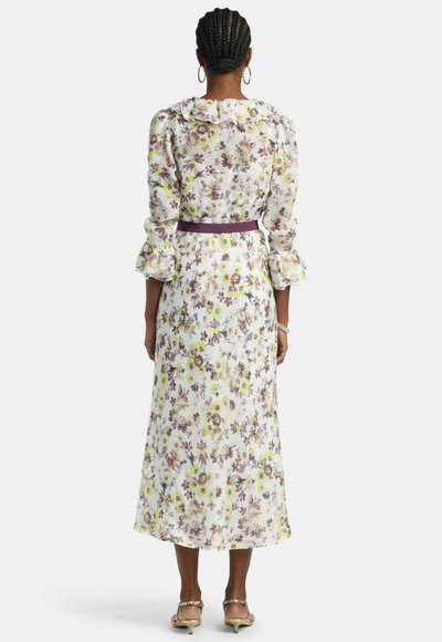 Femeie cu părul împletit, purtând o rochie lungă cu flori, cu mâneci și guler cu volane, curea mov și sandale aurii, stând cu spatele la cameră.