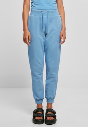 Hellblaue Jogginghose mit elastischen Bündchen und Kordelzugbund. Getragen mit schwarzen Slip-On Sandalen mit drei breiten Riemen.