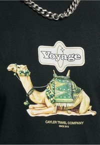 Sort t-shirt med et grafisk print af en kamel prydet med en grøn og mønstret saddel, med teksten "Voyage" og "Cayler Travel Company" nedenunder.