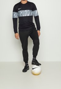 Czarna, długorękawowa koszulka Nike z odblaskowym szarym pasem, zestawiona z czarnymi dresami i czarnymi butami do piłki nożnej, stojąca obok białej i złotej piłki nożnej.