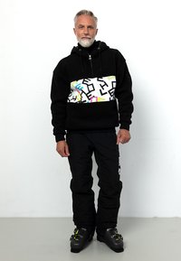 Schwarz mit Kapuze, Sweatshirt mit buntem Grafikmuster und Reißverschluss, kombiniert mit schwarzen Schneehosen und schwarzen Skischuhen mit grünen Akzenten.