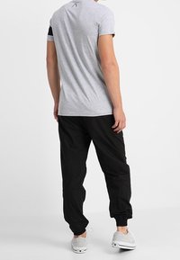 Muž v světle šedém tričku, černých joggingových kalhotách a šedých teniskách stojící na jednobarevném bílém pozadí.