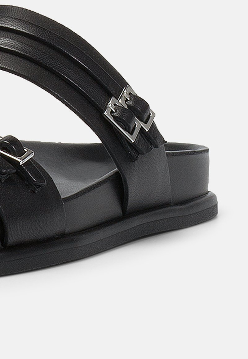 Schwarze Ledersandale mit zwei Riemen, silbernen Schnallen und einer flachen Gummisohle, die eine glatte Textur und ein minimales Design aufweist.