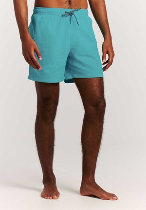 Homme portant un short de bain turquoise avec un cordon noir et blanc, debout pieds nus sur un fond uni.