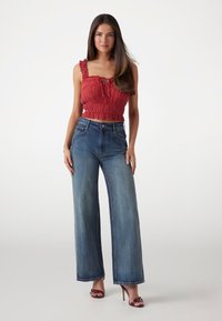 Top court rouge à smocks avec des bretelles à volants, associé à un jean en denim bleu à taille haute et à jambes larges. Modèle portant des talons ouverts marron.
