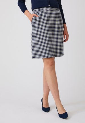 Vrouw die een knielange blauw-wit geruite rok met zakken draagt, een donkerblauwe cardigan en donkerblauwe hoge hakken.
