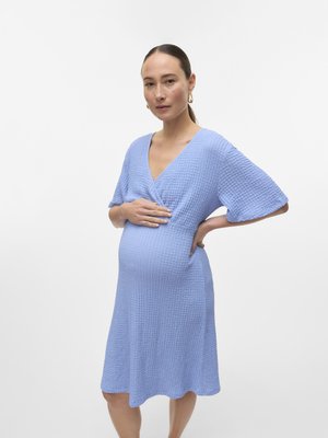 Femme enceinte aux cheveux attachés porte une robe bleu clair texturée, main posée sur le ventre et la hanche, sur un fond blanc uni.
