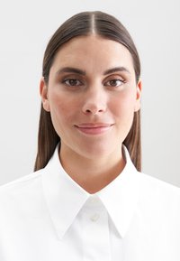 Giovane donna con capelli castani lisci, che indossa una camicia bianca con colletto, che guarda direttamente con un leggero sorriso su uno sfondo chiaro e semplice.
