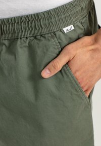 Pantalones cortos de algodón verde con cintura elástica, bolsillos laterales y una pequeña etiqueta de la marca cosida en la cadera. Textura suave, corte relajado.