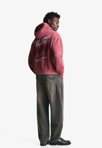 Bordo dukserica s kapuljačom s grafičkim natpisom na leđima, u kombinaciji s suženim sivoj hlačama. Crne cipele upotpunjuju outfit. Glatka, pamučna tekstura.