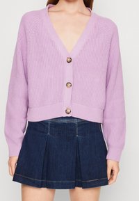 Cardigan en tricot lilas avec un col en V, des manches longues et trois gros boutons ronds. Porté sur une jupe plissée en denim foncé.