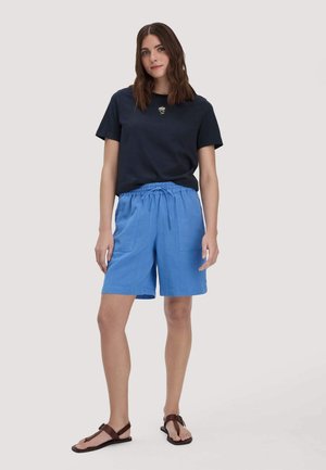 Frau steht vor einfachem Hintergrund, trägt ein schwarzes T-Shirt mit kleinem Blumenmuster, blaue Shorts mit Kordelzug und braune Sandalen.