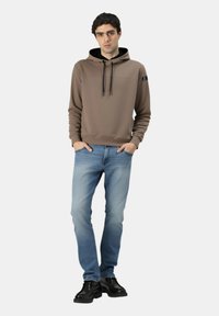 Giovane uomo in piedi con le mani in tasca, indossa una felpa marrone con cappuccio, jeans blu sbiaditi e scarpe di pelle nere su uno sfondo semplice.