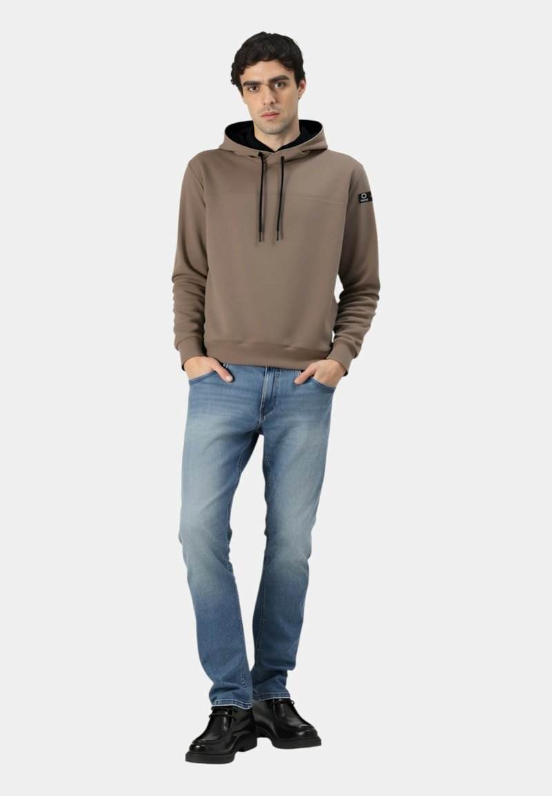 Giovane uomo in piedi con le mani in tasca, indossa una felpa marrone con cappuccio, jeans blu sbiaditi e scarpe di pelle nere su uno sfondo semplice.