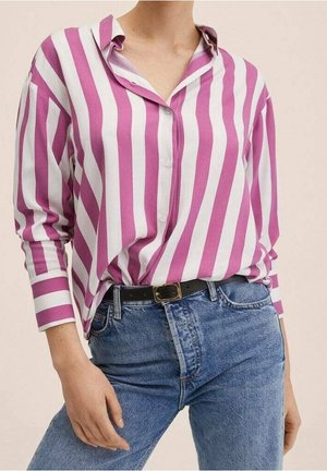 Femme portant une chemise à manches longues boutonnée avec des rayures verticales rose et blanc, rentrée dans un jean bleu taille haute avec une ceinture noire.