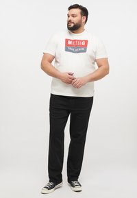 Camiseta blanca con el logo "MUSTANG TRUE DENIM" en rojo y azul, combinada con vaqueros negros y zapatillas negras. Diseño simple y casual, sin adornos.