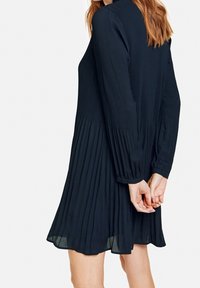 Marinblå plisserad klänning med långa ärmar, rund halsringning och utsvängd nederkant. Tillverkad av lättviktsmaterial, har vertikala plisseringar och manschettärmar.
