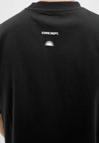 Camiseta negra de cuello redondo, con el texto blanco "CORE DEPT." y un gráfico de sol en la parte superior de la espalda. Hecha de una tela suave.
