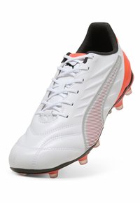 Botte de football blanche avec une surface texturée, présentant une bande argentée et rouge, des accents noirs et une semelle rose avec des crampons moulés.