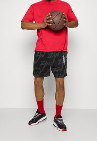 Camiseta de algodón roja, pantalones cortos negros con un audaz patrón gris, calcetines rojos y zapatillas negras con cordones blancos y detalles rojos. Sosteniendo un balón de baloncesto.