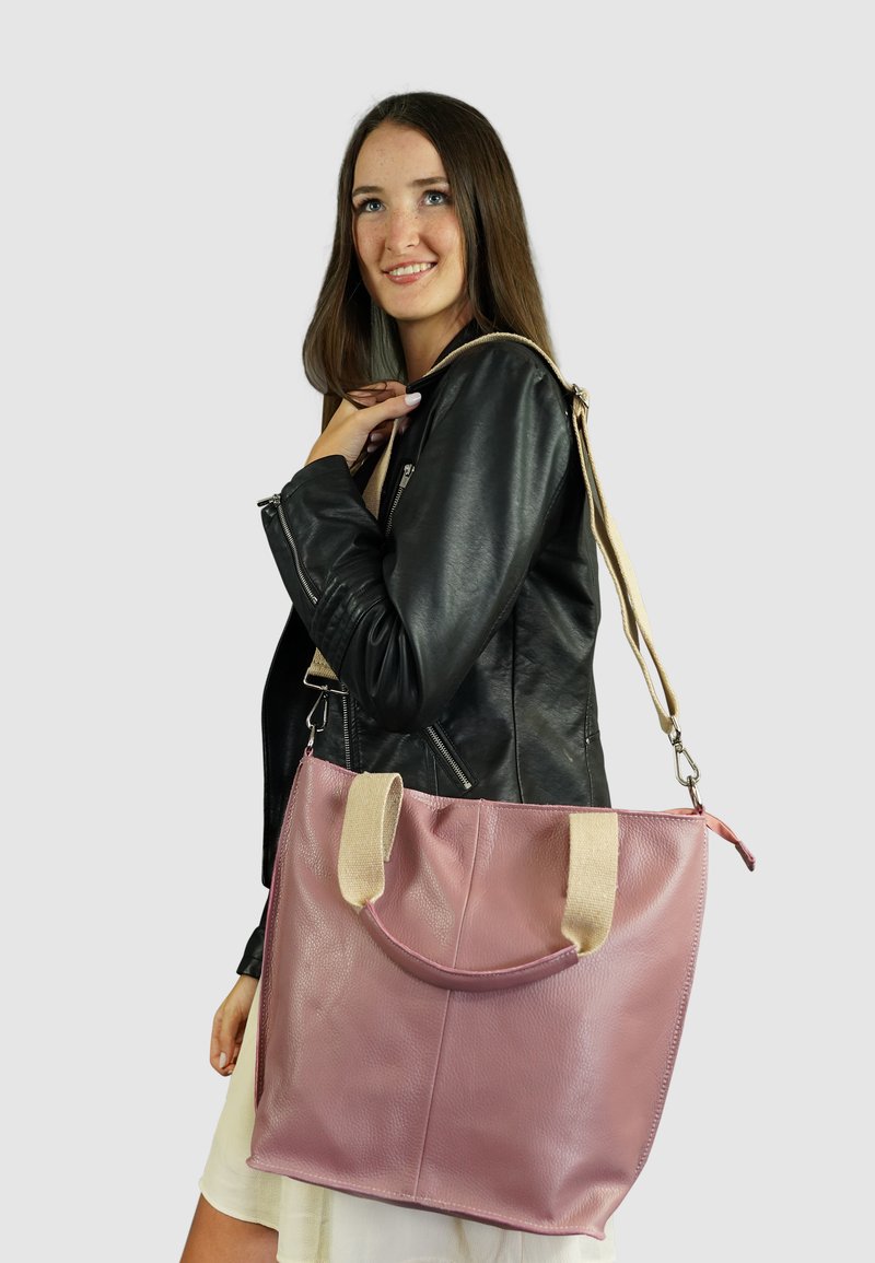 Grote roze leren tote bag met beige stoffen handvatten, met een gladde textuur en een gebogen opening bovenaan, vastgehouden door een persoon in een zwarte jas.