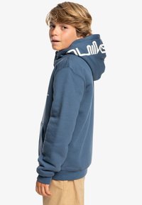 Sweat à capuche bleu avec un grand logo blanc dans le dos, manches longues, poche kangourou et poignets côtelés, porté sur un pantalon beige.