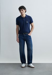 Μπλε ναυτικό πόλο με λογότυπο, υφασμάτινη υφή· συνδυασμένο με μπλε τζιν και λευκά αθλητικά παπούτσια. Άνετη εφαρμογή με κλασικό γιακά και κοντά μανίκια.