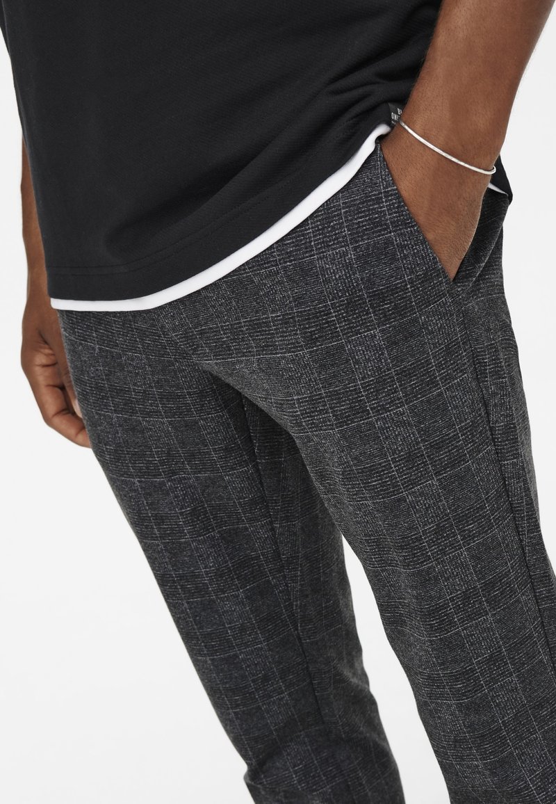 Pantalons à carreaux gris avec un tissu texturé, présentant une coupe droite et des poches latérales. Portés avec une chemise noire superposée en dessous.