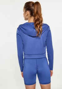 Completo sportivo blu composto da un hoodie corto con cappuccio e pantaloni corti aderenti coordinati. Realizzato in un tessuto liscio e elasticizzato con una leggera lucentezza.