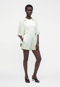 Femme portant un haut blanc sans manches, une chemise légère verte ouverte à manches courtes, un short assorti et des sandales à talons noires, debout avec une main dans la poche.