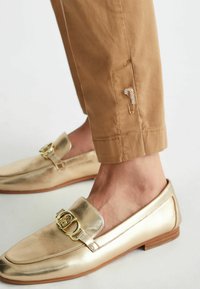 Mocassini dorati con una finitura metallica lucida, caratterizzati da un dettaglio hardware decorativo sulla parte superiore. Indossati con pantaloni beige a vita alta.