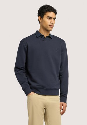 Mann trägt ein marineblaues Sweatshirt mit Rundhalsausschnitt über einem dunklen Hemd und beigen Hosen, steht mit einer Hand in der Tasche.