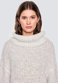 Gruby sweter dziany w jasnym beżu, z szerokim kołnierzem typu cowl i luźnym krojem. Tekstura jest gruba, z otwartym splotem.