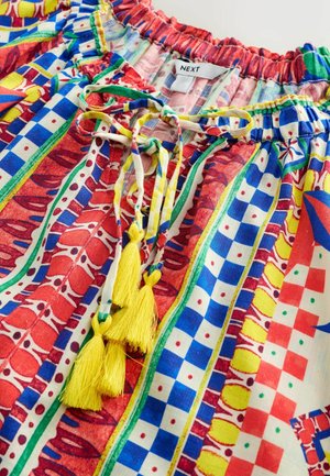 Barevná vzorovaná halenka s červenými, modrými a žlutými akcenty, s nařaseným výstřihem a žlutými střapcovými pásky vpředu.