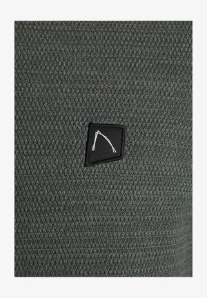 Harmaa neulottu kangas, jossa on teksturoitu kuvio; esittelee mustan kangasmerkin, jossa on valkoinen geometrinen muotoilu kulmassa.