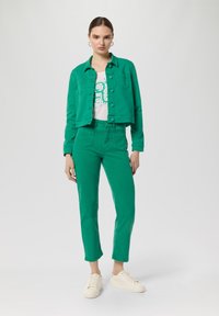 Giacca di denim verde abbinata a pantaloni verdi coordinati. La giacca ha un design cropped e chiusura con bottoni, mentre i pantaloni presentano tasche frontali.