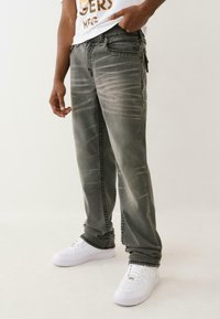 Osoba ubrana w ciemnoszare, wyblakłe dżinsy, białe sneakersy i białą koszulkę z grafiką, stojąca z jedną ręką w kieszeni.