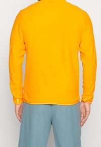 Suéter de forro polar naranja brillante con mangas largas, escote redondeado, puños elásticos y una textura suave. Presenta una costura horizontal en la parte posterior.