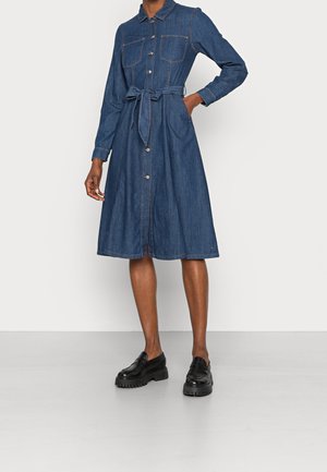 Kobieta ubrana w zapinaną na guziki, długorękawową sukienkę dżinsową z przednimi kieszeniami, zawiązanym paskiem w talii i czarne masywne mokasyny.