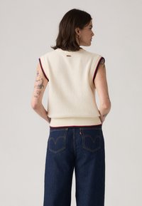 Suéter de punto sin mangas en color crema, con bordes en rayas rojas y azules. Combinado con unos pantalones vaqueros de pierna ancha en azul oscuro con un diseño clásico de bolsillo trasero.