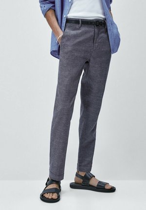 Mann trägt graue Slim-Fit-Hose, schwarzen Gürtel, schwarze Ledersandalen, weißes T-Shirt und blaues Hemd mit Knöpfen, die Hände in den Taschen.