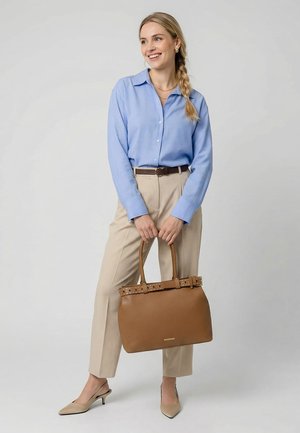 Sieviete ar blondu pinumu, valkājot gaiši zilu blūzi un bēšas bikses, tur lielu brūnu rokassomu, smaida un skatās uz sāniem.