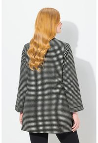 Veste à pois noir et blanc avec une coupe décontractée, des manches longues et une silhouette droite, présentant un tissu texturé et un col rond.