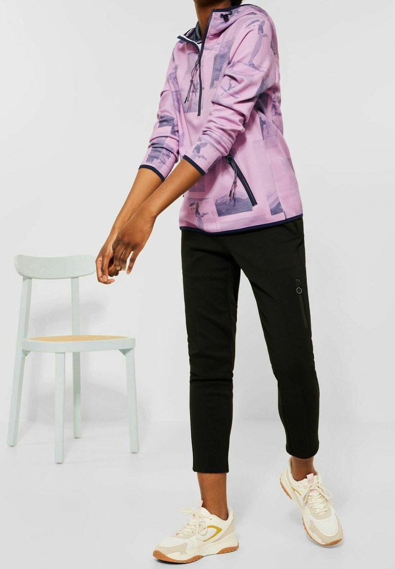 Personne portant un sweat à capuche rose à motifs et un pantalon noir se tient près d'une chaise blanche, portant des baskets blanches et beiges sur un sol gris.