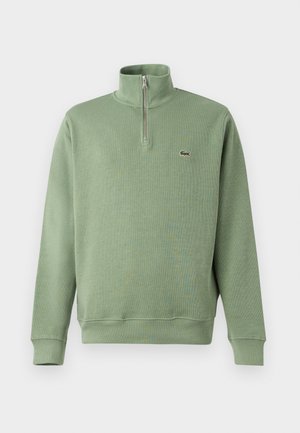 Zelená zip-up mikina s vysokým golierom, rebrovanou textúrou a malým logom Lacoste na ľavej hrudi. Dlhé rukávy a priliehavé manžety.