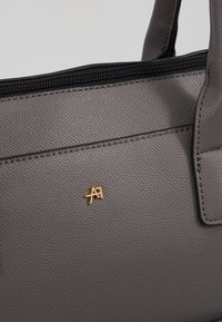 Sac à main gris texturé avec fermeture éclair noire. Accent de logo doré sur le devant. Design structuré avec doubles poignées et détails de couture visibles.