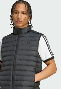 Ung person med flettede hår iført en sort polstret vest over en hvid kortærmet skjorte med sorte striber, der står med den ene hånd i lommen.