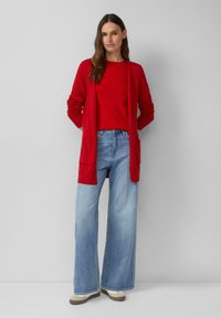 Kobieta stojąca przy szarej ścianie, ubrana w czerwoną kardigan i bluzkę oraz jasnoniebieskie dżinsy z szerokimi nogawkami i beżowe trampki.